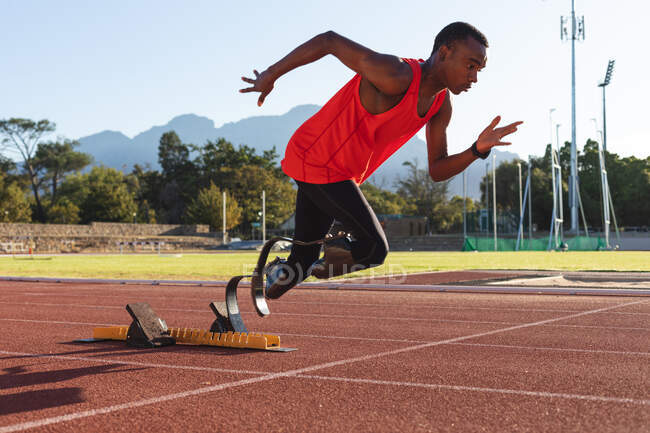 In forma, atleta maschio disabile di razza mista in uno stadio sportivo all'aperto, partendo sprint da blocchi di partenza in pista con lame da corsa. Disabilità atletica allenamento sportivo. — Foto stock