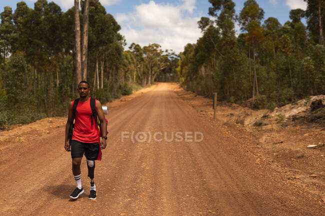 Athlète masculin handicapé de race mixte en forme avec une jambe prothétique, profitant de son temps lors d'un voyage, d'une randonnée, d'une marche sur un chemin de terre dans une forêt. Mode de vie actif avec handicap. — Photo de stock