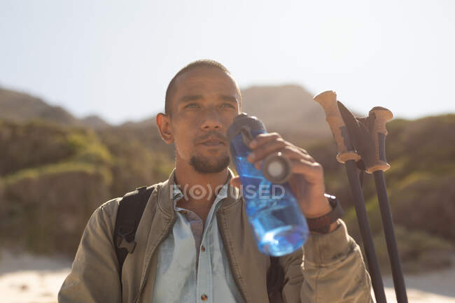 A fit, mixed race male athlete, enjoying his time on a trip to the mountains, hiking with sticks, drinking water beach, resting on the beach by the sea. Active lifestyle Nordic walking. — Stock Photo