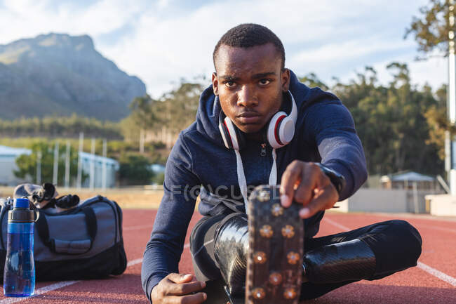 Fit, mixed race disabled male athlete at an outdoor sports stadium, with gym bag and water bottle stretching on race track wearing running blades. Disability athletics sport training. — Stock Photo