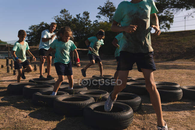 Группа кавказских детей в учебном лагере в солнечный день, в зеленых футболках и черных шортах, бегущих по шинам на полосе препятствий, с кавказским тренером на заднем плане — стоковое фото
