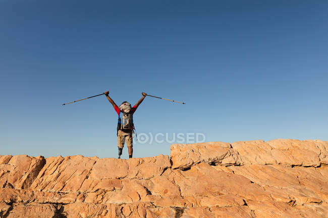 Fit, disabled mixed race male athlete with prosthetic leg, enjoying his time on a trip to the mountains, hiking on the rocks with his arms above his head with sticks. Active lifestyle with disability. — Stock Photo