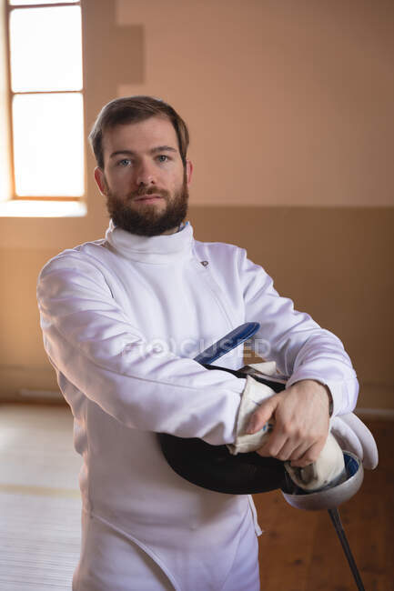 Portrait of Caucasian sportsman wearing protective fencing outfit during a fencing training session, looking at camera, holding an epee and a mask. Fencers training at a gym. — Stock Photo