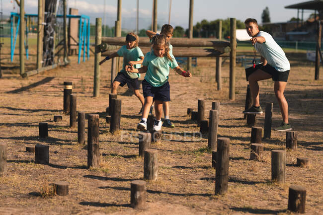 A Caucasian male fitness coach instructing a group of Caucasian boys and girls wearing green t shirts and black shorts at a boot camp on a sunny day, balancing and walking on logs of different height — Stock Photo