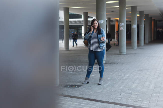 Kurvige Kaukasierin, die tagsüber auf den Straßen der Stadt unterwegs ist, lächelt, gestikuliert und mit ihrem Smartphone spricht, im Hintergrund modernes Gebäude — Stockfoto