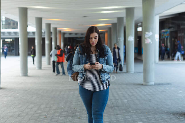 Curvy femme caucasienne dans les rues de la ville pendant la journée, souriant et utilisant son smartphone, marchant avec un bâtiment moderne en arrière-plan — Photo de stock