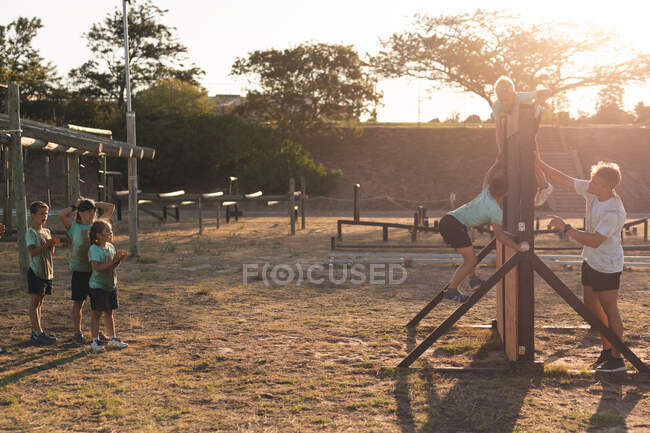 Un entrenador de fitness masculino caucásico que ayuda a un grupo de niños y niñas caucásicos que usan camisetas verdes y pantalones cortos negros en un campamento de entrenamiento en un día soleado, trepando por una valla de madera en una carrera de obstáculos - foto de stock