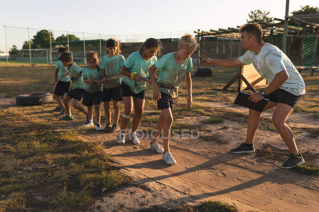 Un entraîneur de fitness masculin caucasien instruisant un groupe de garçons et de filles caucasiens portant des t-shirts verts et des shorts noirs lors d'un camp d'entraînement par une journée ensoleillée, tirant un pneu avec une corde — Photo de stock