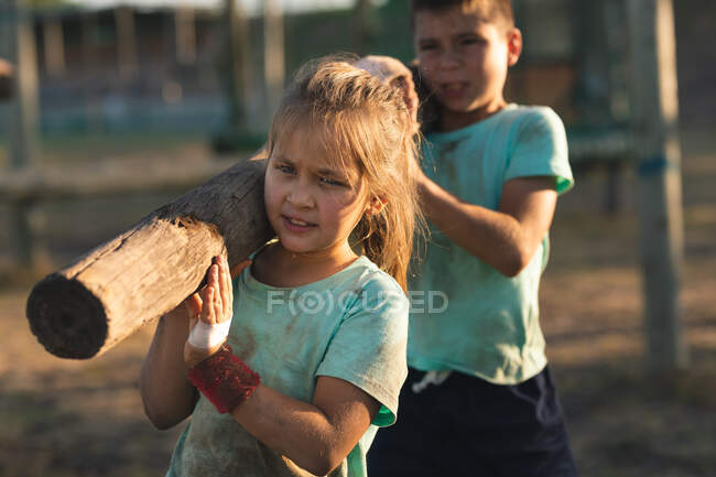 Кавказская девушка и мальчик в грязно-зеленых футболках и черных шортах, несущих бревно вместе на своих плечах во время тренировки в учебном лагере в солнечный день — стоковое фото