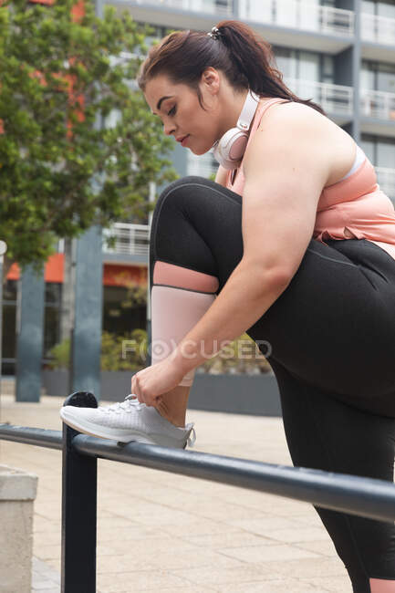 Curvy Caucasian woman with long dark hair wearing sports clothes and headphones exercising in a city, with her foot up on a fence, tying her shoelaces, with modern buildings behind her — Stock Photo