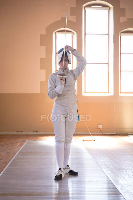 Portrait of Caucasian sportswoman wearing protective fencing outfit during a fencing training session, looking at camera and smiling, holding an epee. Fencers training at a gym. — Stock Photo