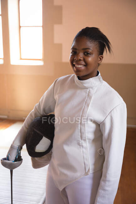 Porträt einer afroamerikanischen Sportlerin, die während eines Fechttrainings Schutzkleidung trägt, in die Kamera blickt und lächelt, Degen und Maske in der Hand hält. Fechter beim Training im Fitnessstudio. — Stockfoto
