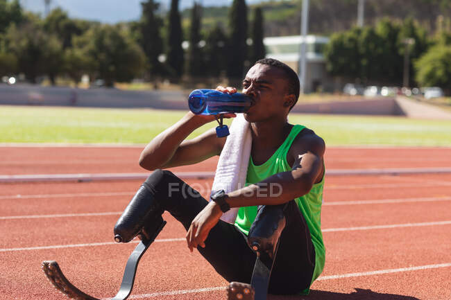 Fit, mixed race disabled male athlete at an outdoor sports stadium, sitting on race track after race drinking water wearing running blades. Disability athletics sport training. — Stock Photo