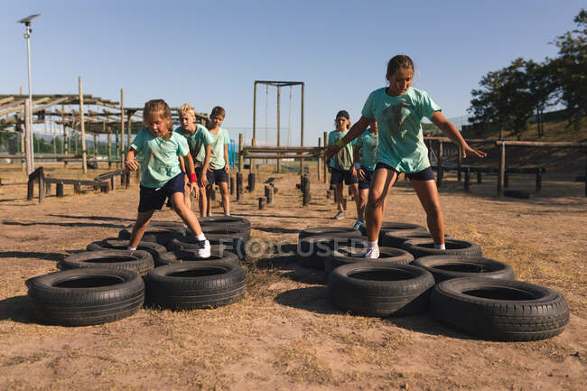 Duas garotas caucasianas em um campo de treinamento em um dia ensolarado, vestindo camisetas verdes e shorts pretos, atravessando pneus em um curso de obstáculo, com outras crianças seguindo-os no fundo — Fotografia de Stock