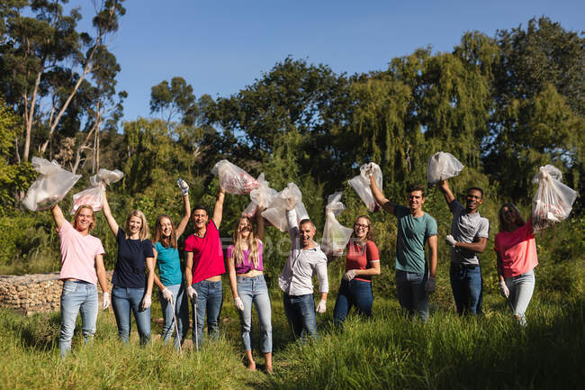 Портрет счастливой многонациональной группы волонтеров-консерваторов, очищающих реку в сельской местности, держащих мусорные мешки в воздухе. Экология и социальная ответственность в сельской местности. — стоковое фото