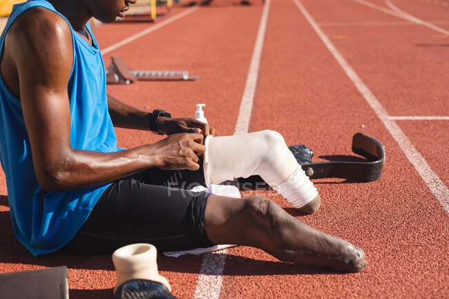 Mid section of fit, mixed race disabled male athlete at an outdoor sports stadium, sitting on race track preparing running blades before workout. Disability athletics sport training. — Stock Photo