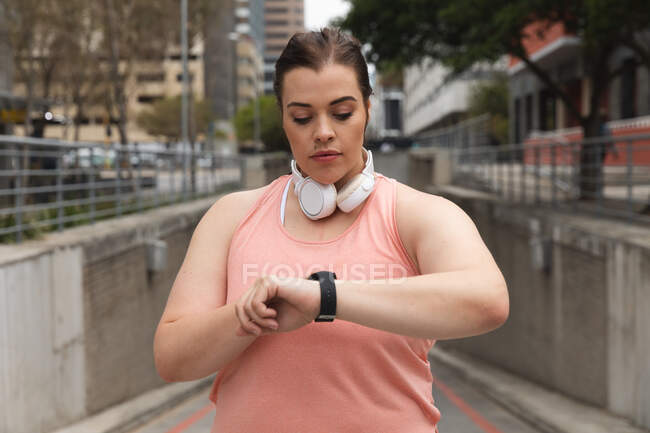 Curvy Caucasian woman with long dark hair wearing sports clothes and headphones exercising in a city, checking her smartwatch, with modern buildings behind her — Stock Photo