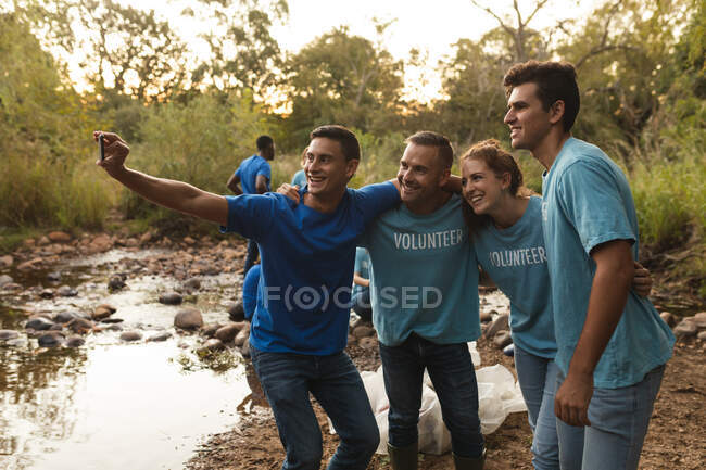 Grupo multiétnico de voluntarios felices de conservación limpiando el río en el campo, tomando selfie con smartphone. Ecología y responsabilidad social en el medio rural. - foto de stock