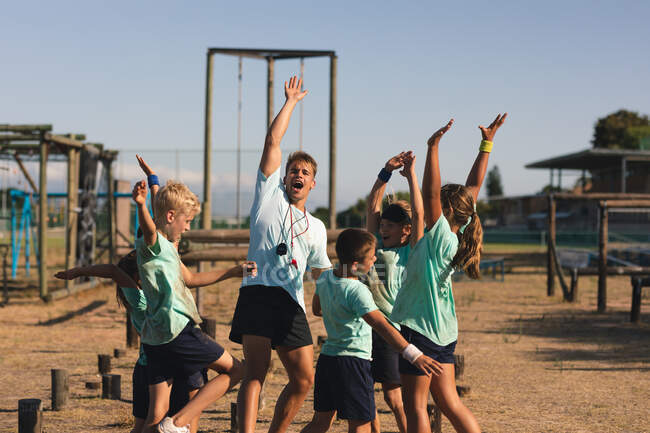 A group of Caucasian boys and girls and a Caucasian male fitness coach at a boot camp on a sunny day, standing and celebrating, shouting with arms raised in the air at the end of an obstacle course — Stock Photo
