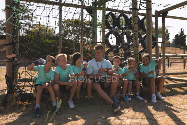 Un groupe de garçons et de filles caucasiens assis avec un entraîneur de conditionnement physique masculin caucasien se reposer, boire des bouteilles d'eau et parler à un camp de démarrage par une journée ensoleillée — Photo de stock