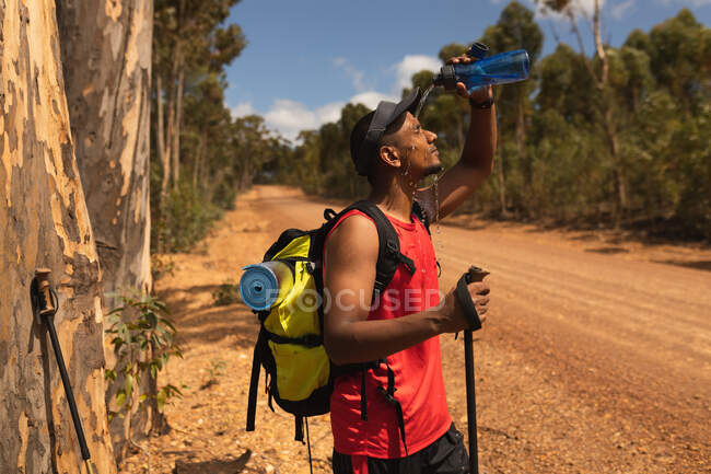A fit, disabled mixed race male athlete with prosthetic leg, enjoying his time on a trip, hiking, standing on a dirt road in a forest, pouring water on himself. Active lifestyle with disability. — Stock Photo