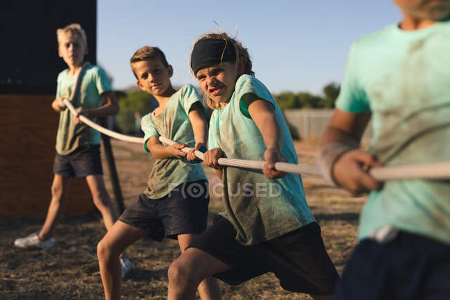 Eine Gruppe kaukasischer Jungen und Mädchen in schlammgrünen T-Shirts und schwarzen Shorts, die bei einem Tauziehen in einem Bootcamp an einem sonnigen Tag ein Seil zusammenziehen — Stockfoto