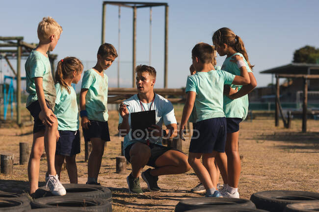 A group of Caucasian boys and girls listening to instructions from a Caucasian male fitness coach at a boot camp on a sunny day, standing and paying attention to him while he squats down and explains — Stock Photo