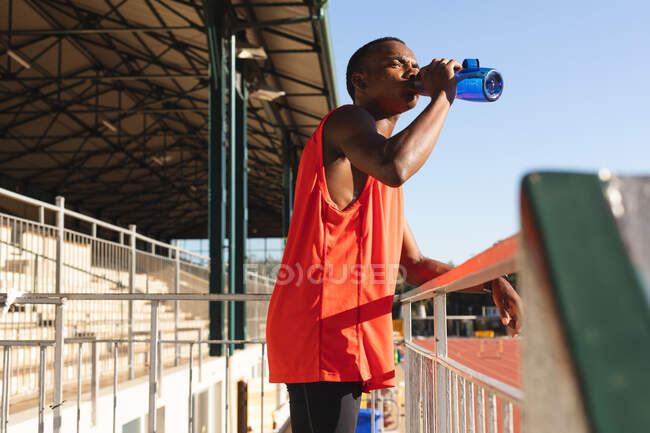 Ajuste, atleta masculino de raza mixta en un estadio de deportes al aire libre, descansando y bebiendo de botella de agua de pie en las gradas. Atletismo entrenamiento deportivo. - foto de stock