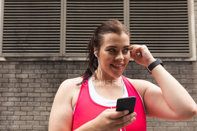 Lächelnde kurvige kaukasische Frau mit langen dunklen Haaren, die in Sportkleidung in einer Stadt trainiert, ihr Smartphone mit Kopfhörern benutzt, im Hintergrund eine Ziegelwand — Stockfoto