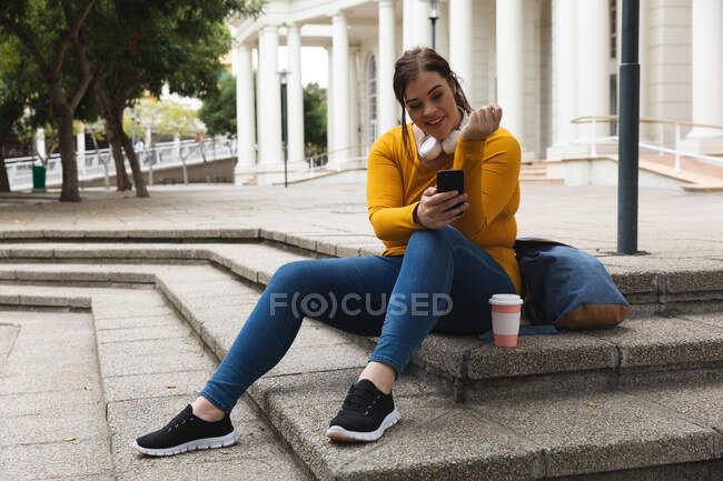 Mujer curvilínea caucásica por las calles de la ciudad durante el día, sentada en escalones con un café, sonriendo y usando su teléfono inteligente usando auriculares con un edificio histórico en el fondo - foto de stock