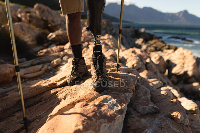 Fit, disabled male athlete with prosthetic leg, enjoying his time on a trip to the mountains, hiking with sticks on the rocks by the sea. Active lifestyle with disability. — Stock Photo