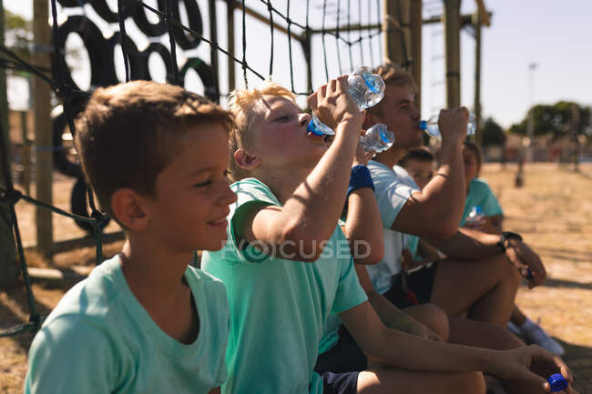 Un grupo de chicos caucásicos sentados con un entrenador de fitness masculino caucásico descansando, bebiendo botellas de agua y sonriendo en un campamento de entrenamiento en un día soleado - foto de stock