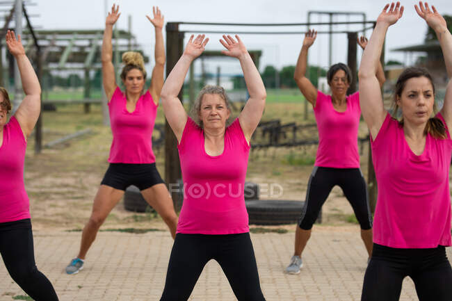 Gruppo multietnico di donne che indossano tutte magliette rosa in una sessione di allenamento di boot camp, esercitandosi, facendo salti jack. Esercizio di gruppo all'aperto, divertente sfida sana. — Foto stock