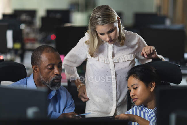 Grupo multiétnico de compañeros de trabajo que trabajan hasta tarde en la noche en una oficina moderna, sentados en un escritorio, usando una tableta y discutiendo su trabajo. - foto de stock