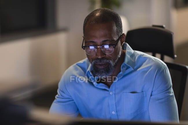 Афроамериканський бізнесмен працює пізно ввечері в сучасному офісі, сидячи за столом, за допомогою настільного комп 