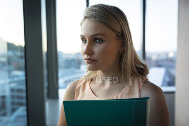Donna d'affari caucasica che lavora fino a tardi la sera in un ufficio moderno, tiene una cartella, guarda attraverso una finestra e pensa:. — Foto stock