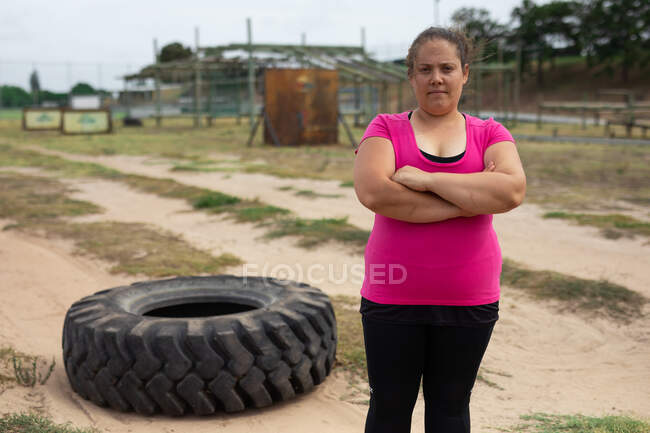 Portrait d'une femme confiante de race mixte dans un camp d'entraînement, portant un t-shirt rose avec les bras croisés, un pneu en arrière-plan. Exercice de groupe en plein air, défi sain amusant. — Photo de stock