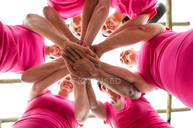 Grupo multiétnico de mujeres que usan camisetas rosas en una sesión de entrenamiento de campo de entrenamiento, ejercitándose, motivándose mutuamente y apilándose las manos. Ejercicio en grupo al aire libre, divertido desafío saludable. - foto de stock