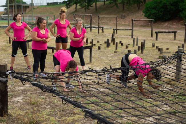 Grupo multiétnico de mujeres todas con camisetas rosas en una sesión de entrenamiento de campo de entrenamiento, haciendo ejercicio, arrastrándose bajo una red. Ejercicio en grupo al aire libre, divertido desafío saludable. - foto de stock