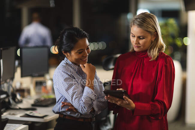 Asian and Caucasian businesswomen working late in the evening in a modern office, using a tablet computer and discussing their work. — Stock Photo