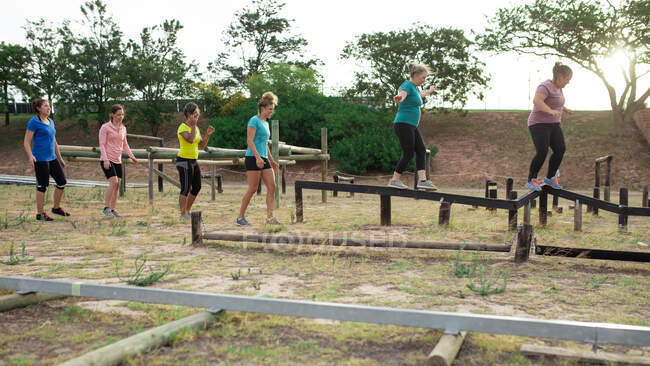 Groupe multi-ethnique de femmes portant toutes des t-shirts colorés lors d'une session d'entraînement de camp d'entraînement, d'exercice, d'équilibrage, de marche sur une poutre. Exercice de groupe en plein air, défi sain amusant. — Photo de stock