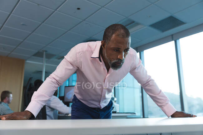 Афроамериканский бизнесмен, работающий допоздна в современном офисе, опираясь на стол, со своими коллегами на заднем плане. — стоковое фото