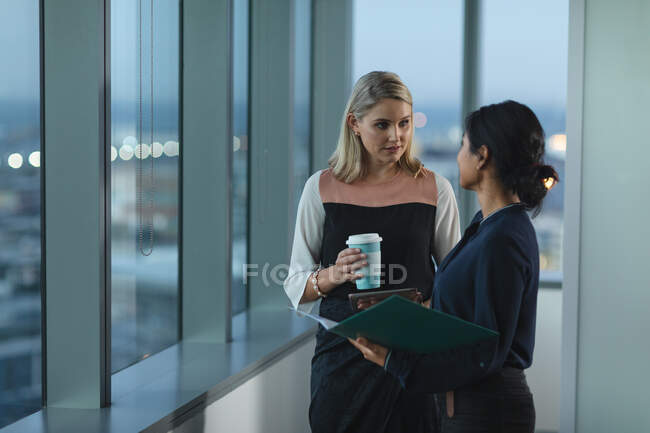Empresarias asiáticas y caucásicas trabajando hasta tarde en una oficina moderna, de pie junto a una ventana y discutiendo su trabajo. - foto de stock