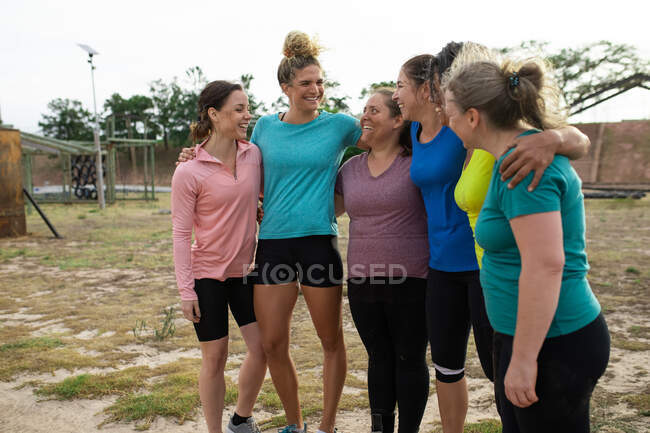 Grupo multi-étnico de mulheres todas vestindo camisetas coloridas em uma sessão de treinamento de campo de treinamento, exercitando-se, motivando umas às outras, abraçando. Exercício de grupo ao ar livre, desafio saudável divertido. — Fotografia de Stock