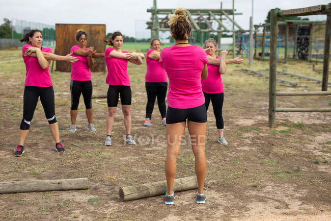 Groupe multi-ethnique de femmes portant toutes des t-shirts roses lors d'une séance d'entraînement dans un camp d'entraînement, faisant de l'exercice, étirant les bras et le canapé les motivant. Exercice de groupe en plein air, défi sain amusant. — Photo de stock