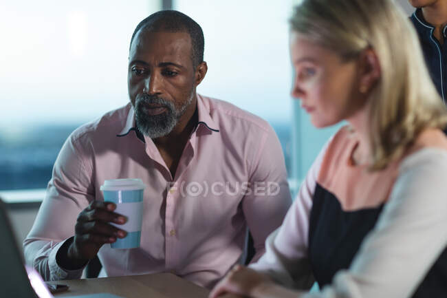 Multi-ethnic group of coworkers working late in the evening in a modern office, sitting at a desk, using a laptop computer and discussing their work. — Stock Photo