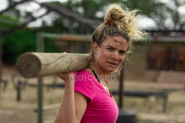 Retrato de uma mulher caucasiana vestindo camiseta rosa em uma sessão de treinamento de campo de treinamento, exercitando-se, carregando um log em suas costas. Exercício de grupo ao ar livre, desafio saudável divertido. — Fotografia de Stock