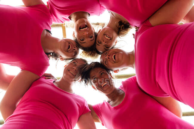 Gruppo multietnico di donne che indossano tutte magliette rosa durante una sessione di allenamento al campo di addestramento, esercitandosi, motivando, abbracciando in gruppo, sorridendo. Esercizio di gruppo all'aperto, divertente sfida sana. — Foto stock