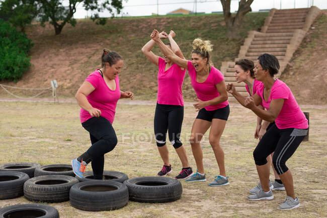 Groupe multi-ethnique de femmes portant toutes des t-shirts roses lors d'une séance d'entraînement dans un camp d'entraînement, faisant de l'exercice, traversant des pneus. Exercice de groupe en plein air, défi sain amusant. — Photo de stock