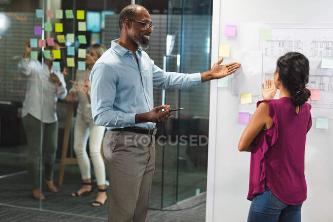 African American businessman and Asian businesswoman working late in the evening in a modern office, writing on a moodboard and brainstorming. — Stock Photo